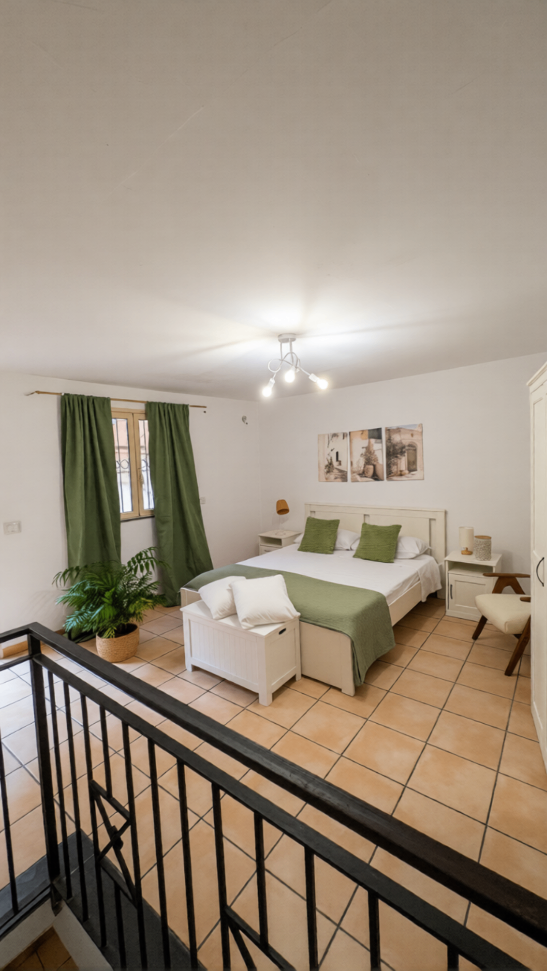 Vista d'insieme della camera matrimoniale al piano superiore con tende verdi, finestra sul vicolo storico e cassapanca ai piedi del letto